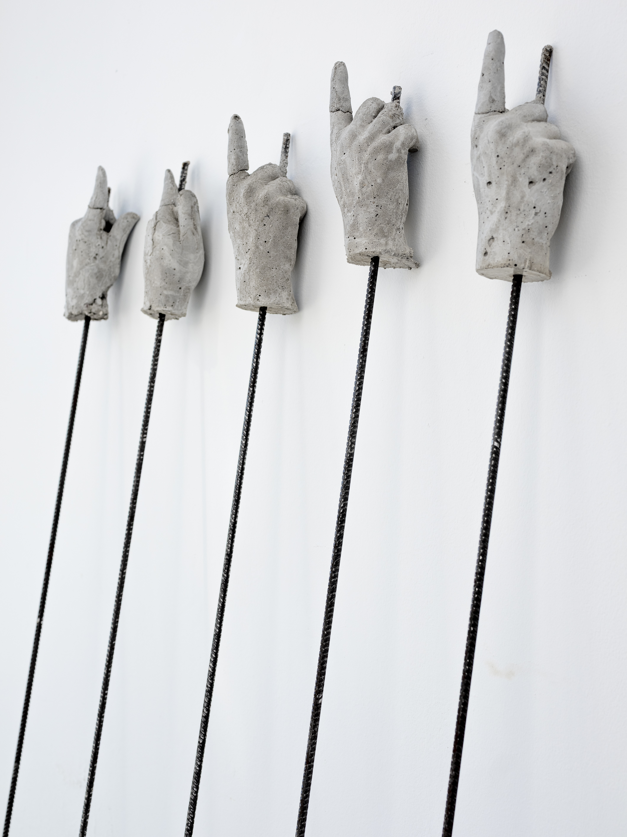 Fünf in Beton gegossene Hände mit der Geste des ausgestreckten Zeigefingers sind auf Metallstäben montiert und lehnen an der Wand im Ausstellungsraum.