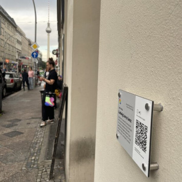 Eine Aufnahme von einen QR-Schild auf der Rosenthaler Straße in Berlin Mitte. Im Hintergrund wird in der Häuserschlucht der Fernsehturm erkennbar, im Fokus ist der an die Wand geschraubte QR-Code.