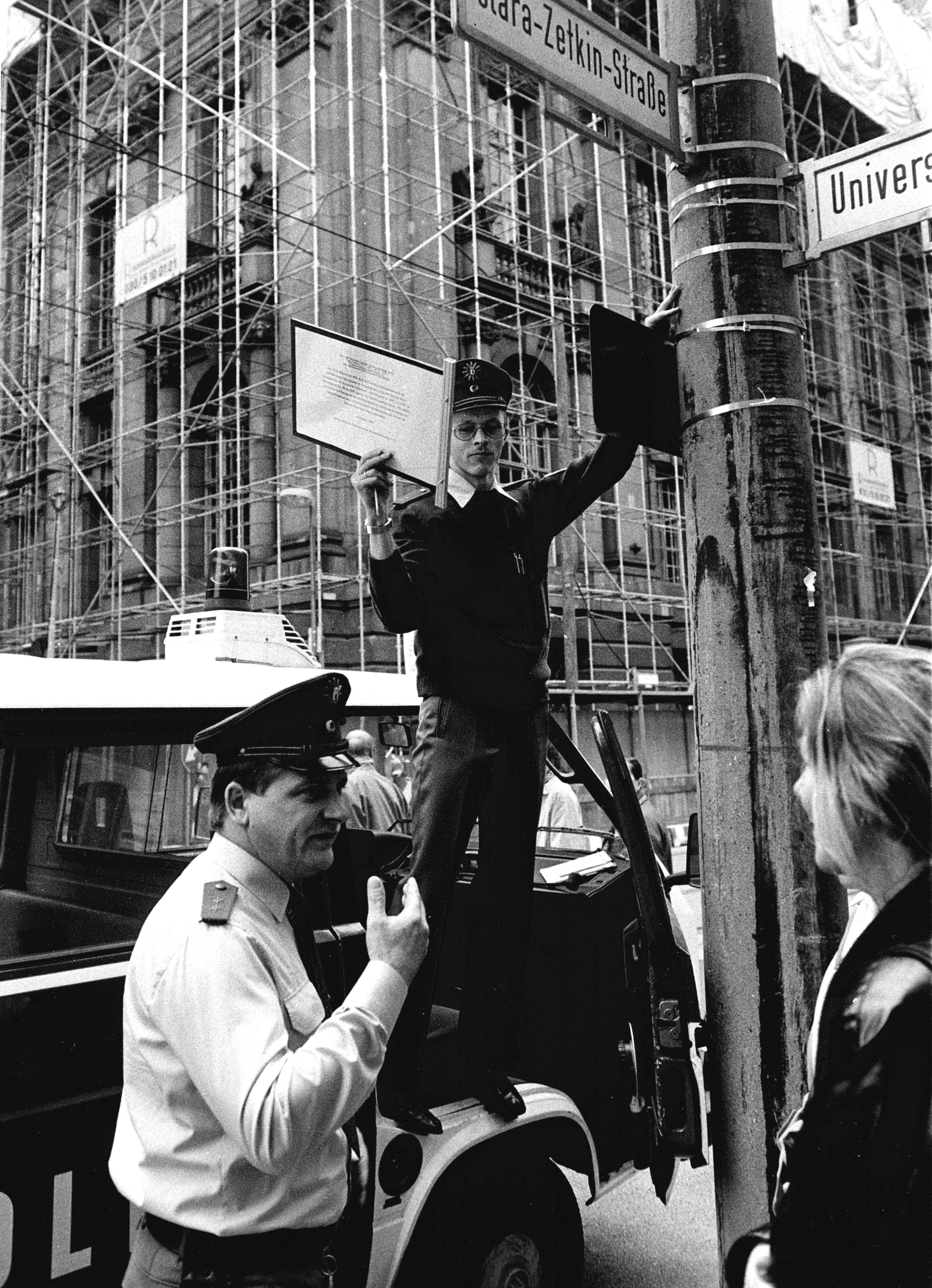 Schwarz-Weiß-Fotografie: Ein Polizist entfernt ein zusätzlich angebrachtes Straßenschild des aktiven Museums, während ein weiterer Beamter im Gespräch mit einer Frau steht.